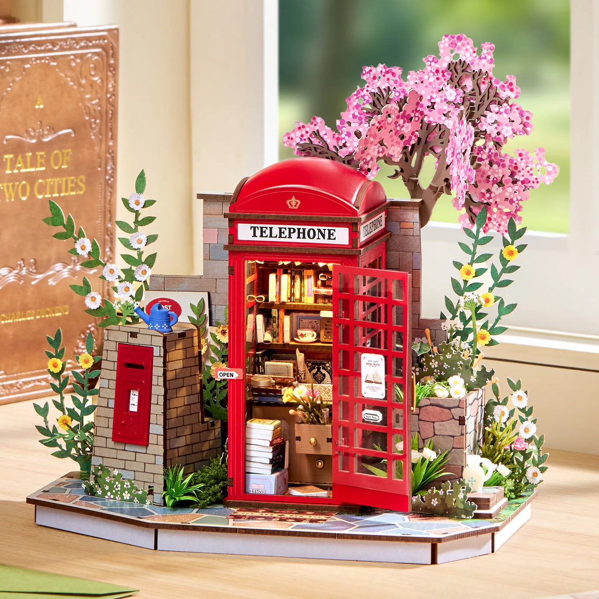 Miniatur-Telefonzelle mit Büchern, umgeben von Blumen und einem Kirschblütenbaum