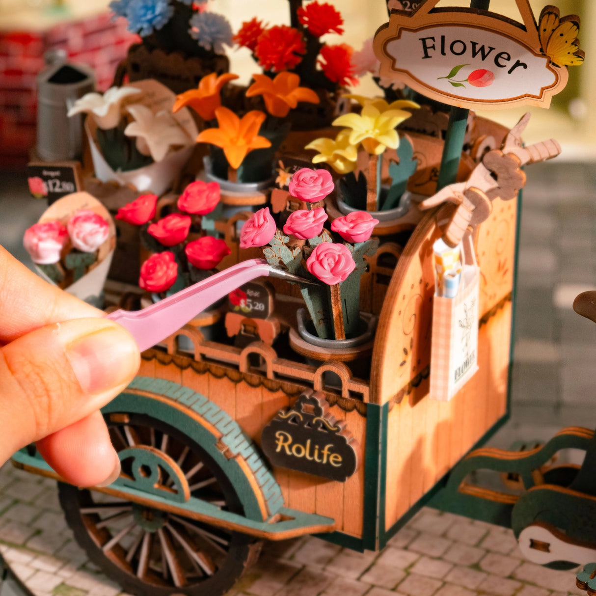 Nahaufnahme eines Miniatur-Blumenwagens mit einer Hand, die eine rosa Pinzette hält und eine rosa Rose platziert