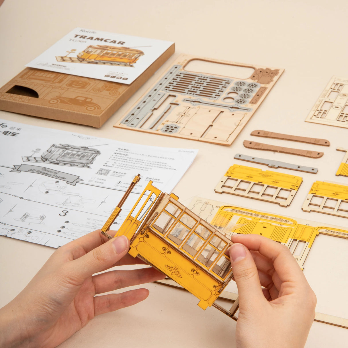 Hände beim Zusammenbau eines gelben 3D Holzpuzzles einer Straßenbahn mit Anleitung und Bauteilen