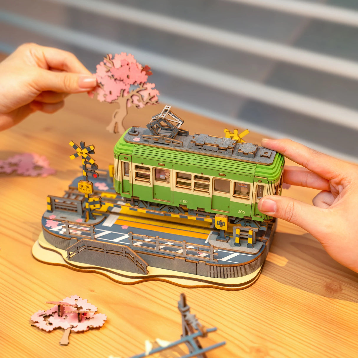 Zwei Hände setzen ein hölzernes Modell einer grünen Straßenbahn mit Kirschblütenbaum zusammen