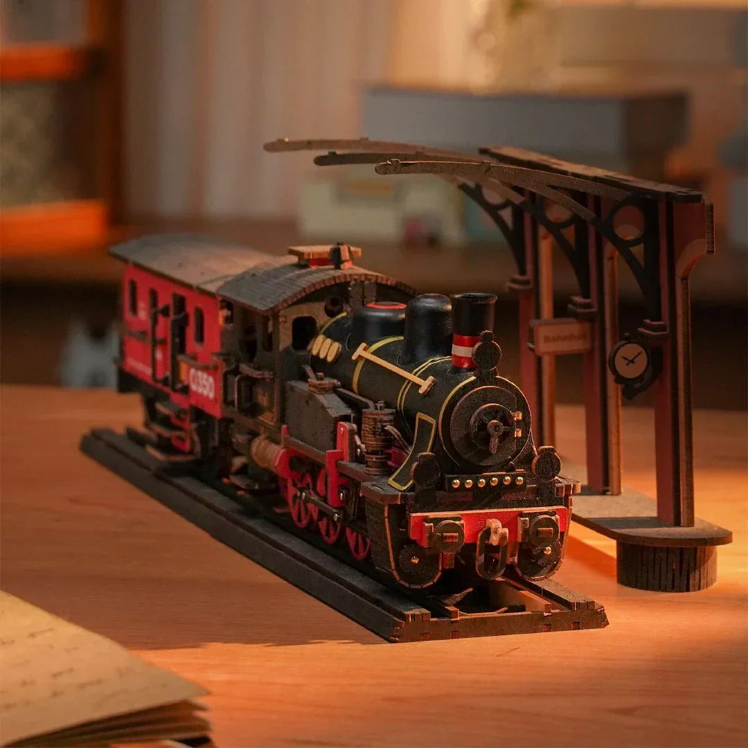 Modell einer schwarzen Dampflokomotive mit roten Rädern und einem roten Waggon auf Holzschienen neben einem Bahnhofsgebäude
