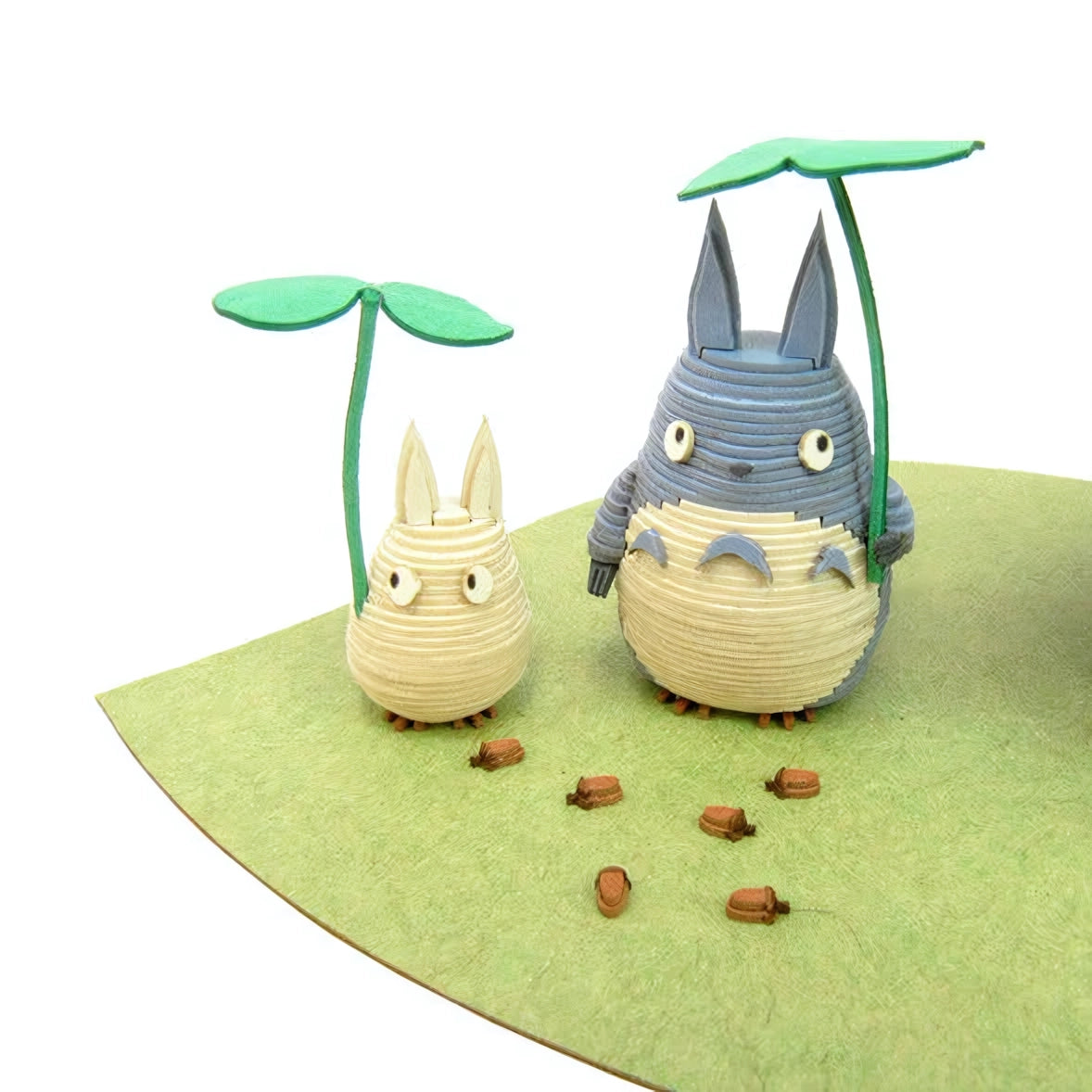 Zwei Totoro-Figuren aus Papier auf grünem Grund, eine hält ein Blatt als Regenschirm