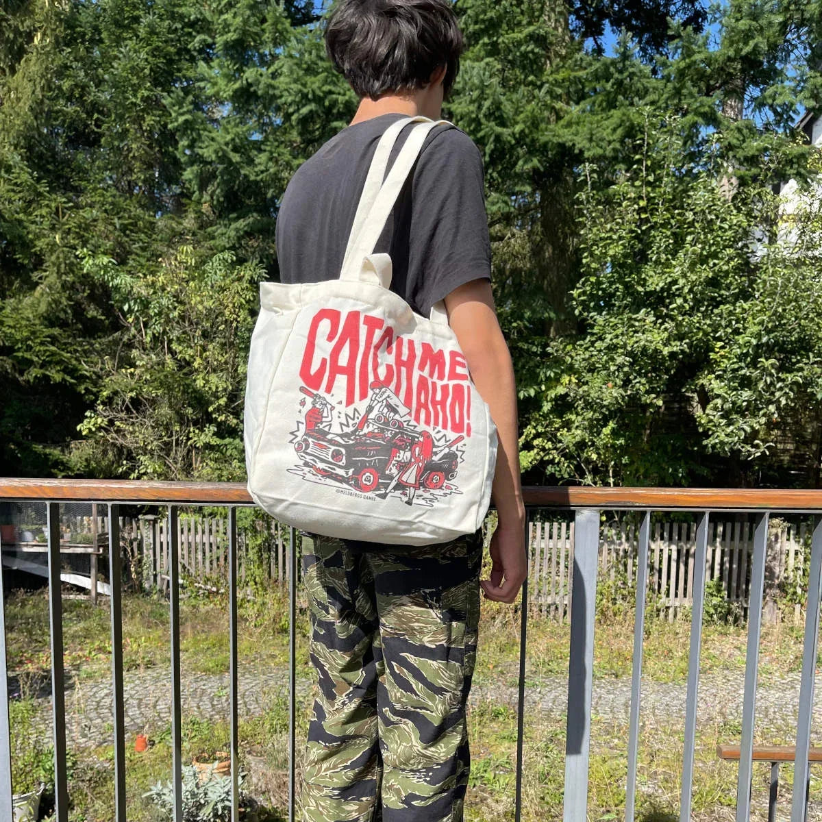 Rückansicht einer Person mit schwarzem T-Shirt, Camouflage-Hose und einer weißen Tragetasche mit rotem Aufdruck