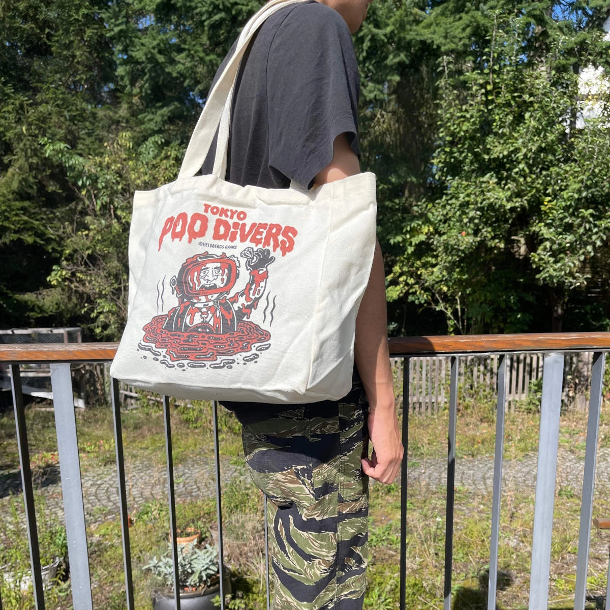 Person trägt eine weiße Stofftasche mit rotem Tokyo Pop Divers Logo und Camouflage-Hose